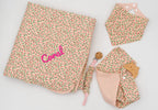 Conjunto rosa com manta de flores e o nome Coral bordado, babete bandana, fralda de musselina rosa com mordedor de madeira e prende-chupetas.
