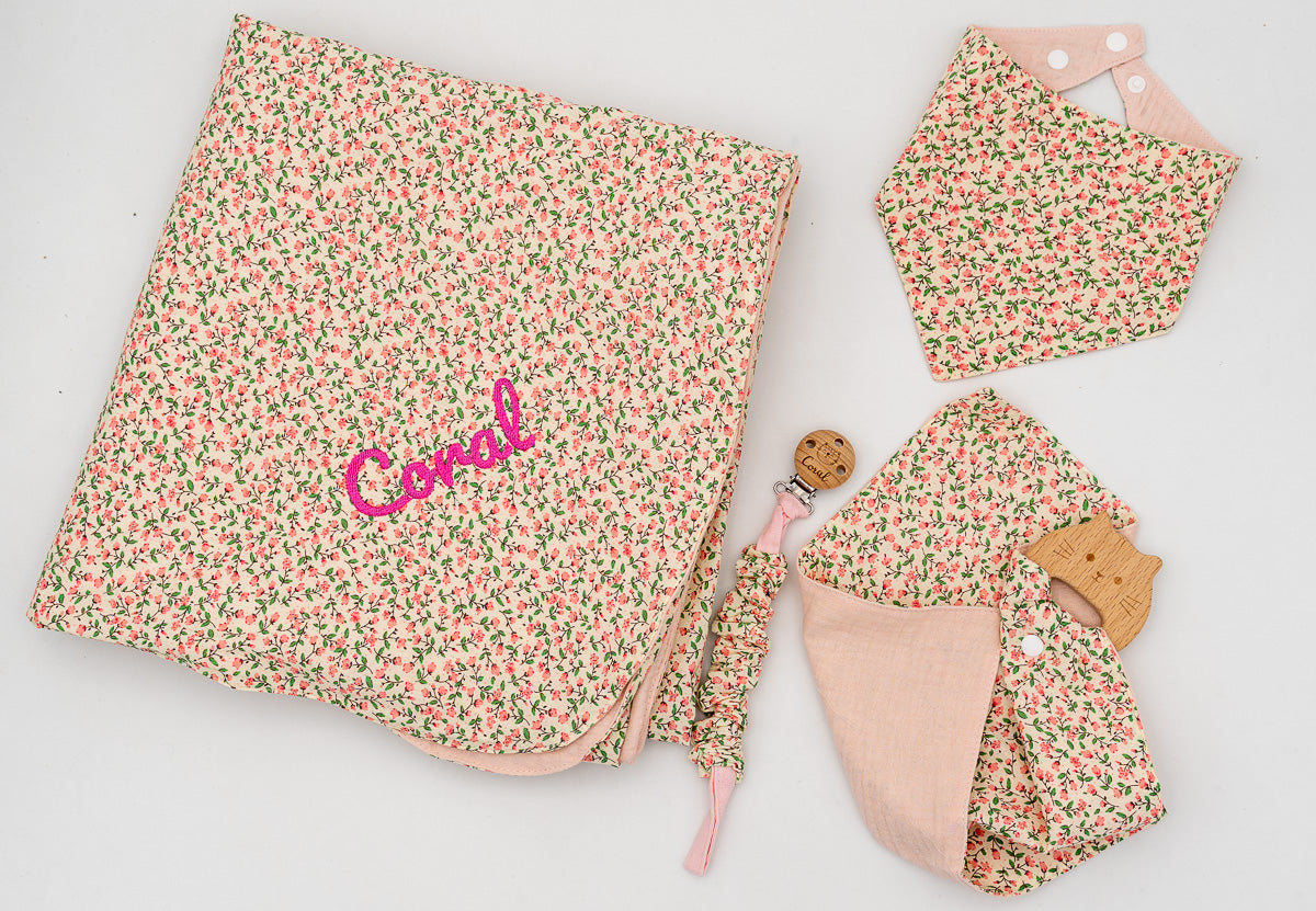 Conjunto rosa com manta de flores e o nome Coral bordado, babete bandana, fralda de musselina rosa com mordedor de madeira e prende-chupetas.