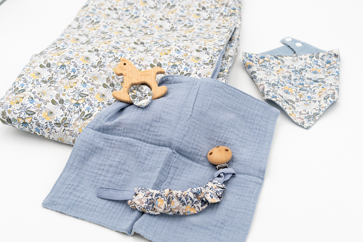 Conjunto azul com manta de flores, babete bandana, fralda de musselina azul com mordedor de madeira em forma de cavalinho e prende-chupetas com mola de madeira.