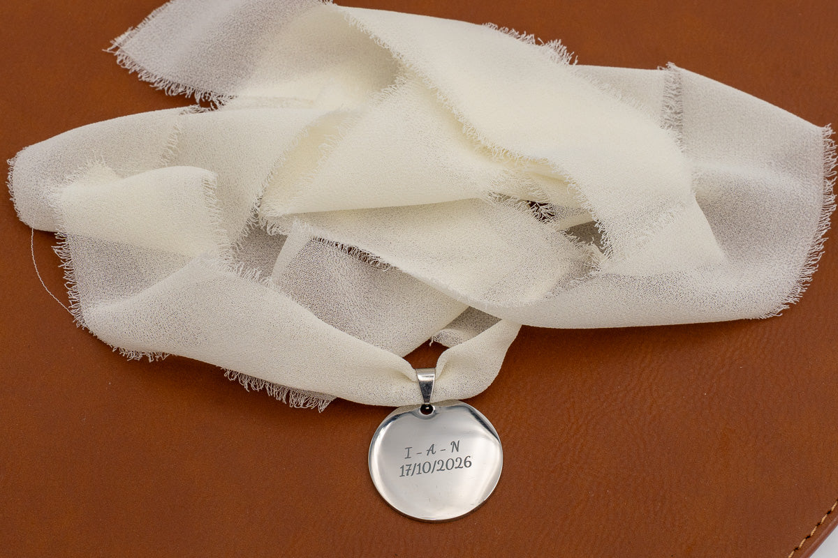 Medalha prateada com iniciais e data gravadas sobre uma fita de chiffon marfim