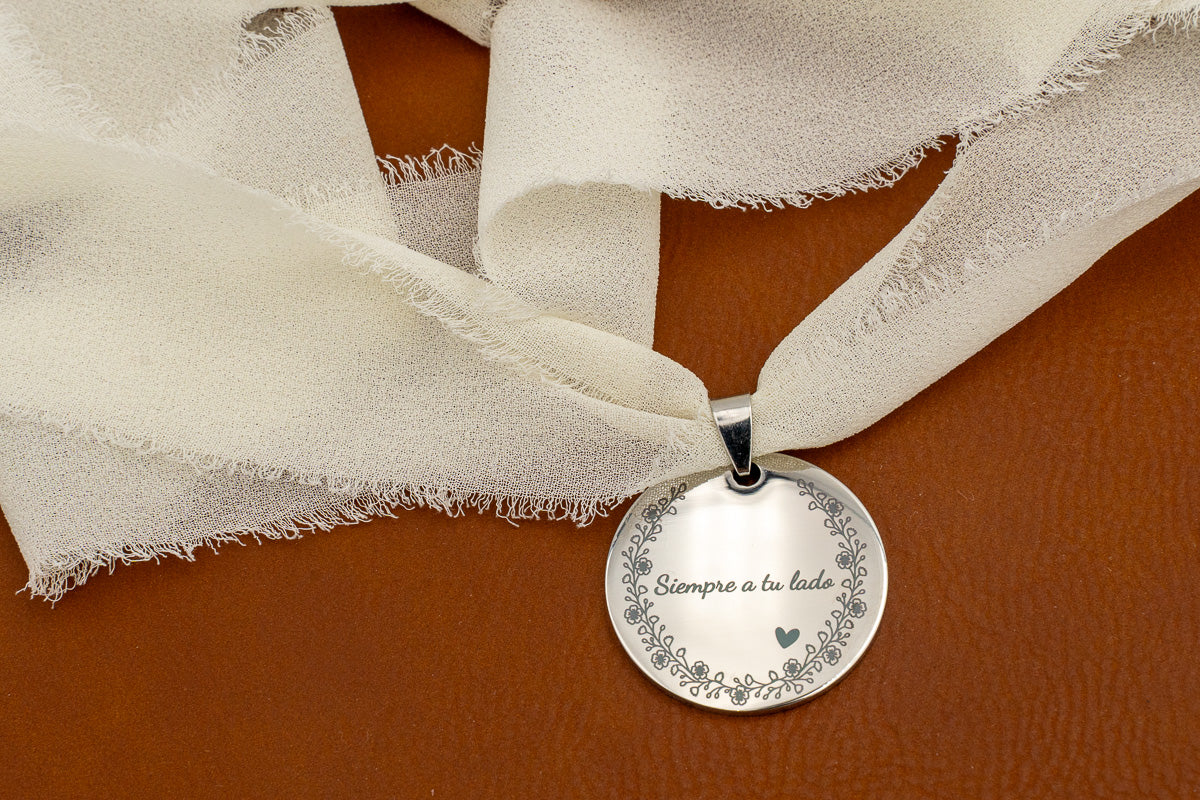 Medalha de aço espelhado com a frase «Sempre ao teu lado» e orla floral, atada com fita de chiffon marfim