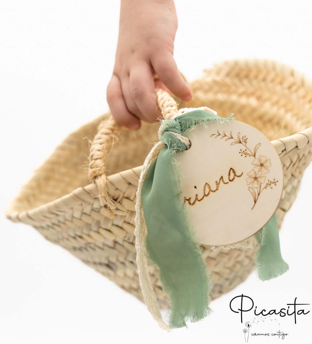Mano infantil sosteniendo un cesto de palma con lazo verde y etiqueta de madera con nombre.
