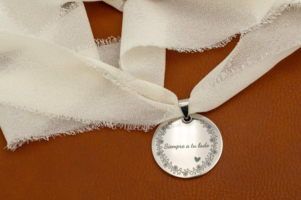 Medalha de aço espelhado com a frase «Sempre ao teu lado» e orla floral, atada com fita de chiffon marfim