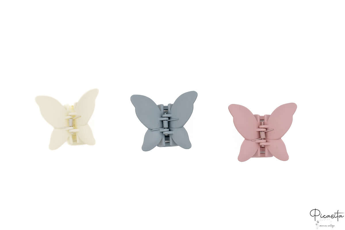 Tres pinzas de pelo con forma de mariposa en colores crema, azul grisáceo y rosa sobre fondo blanco.